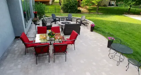 Spacious backyard patio with paver stones featuring outdoor dining set with red cushioned chairs, fire pit seating area with Adirondack chairs, and decorative planters overlooking green lawn